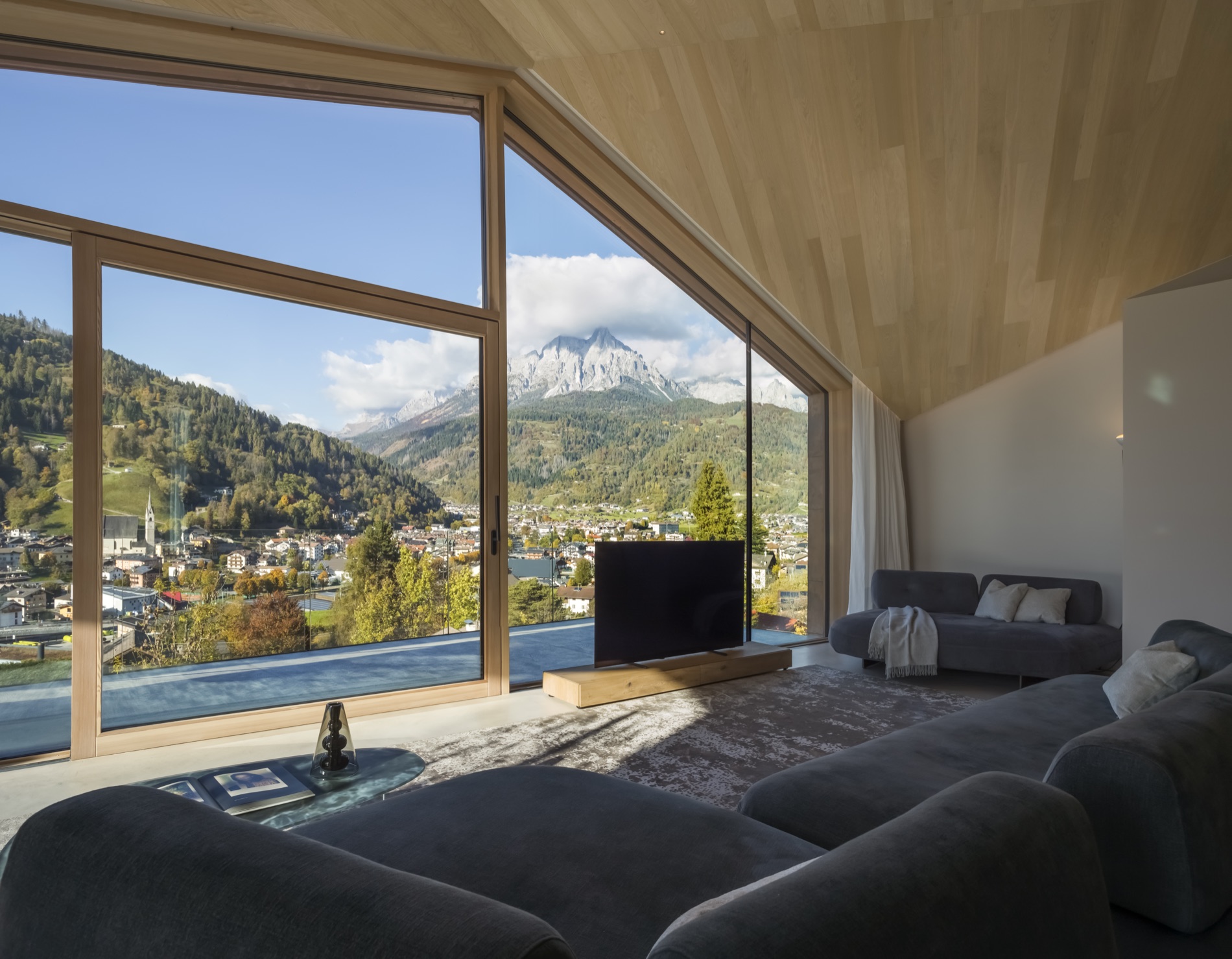 Le séjour panoramique de Casa LAGO Dolomiti, avec de grandes baies ouvertes sur le village de Primiero et les montagnes