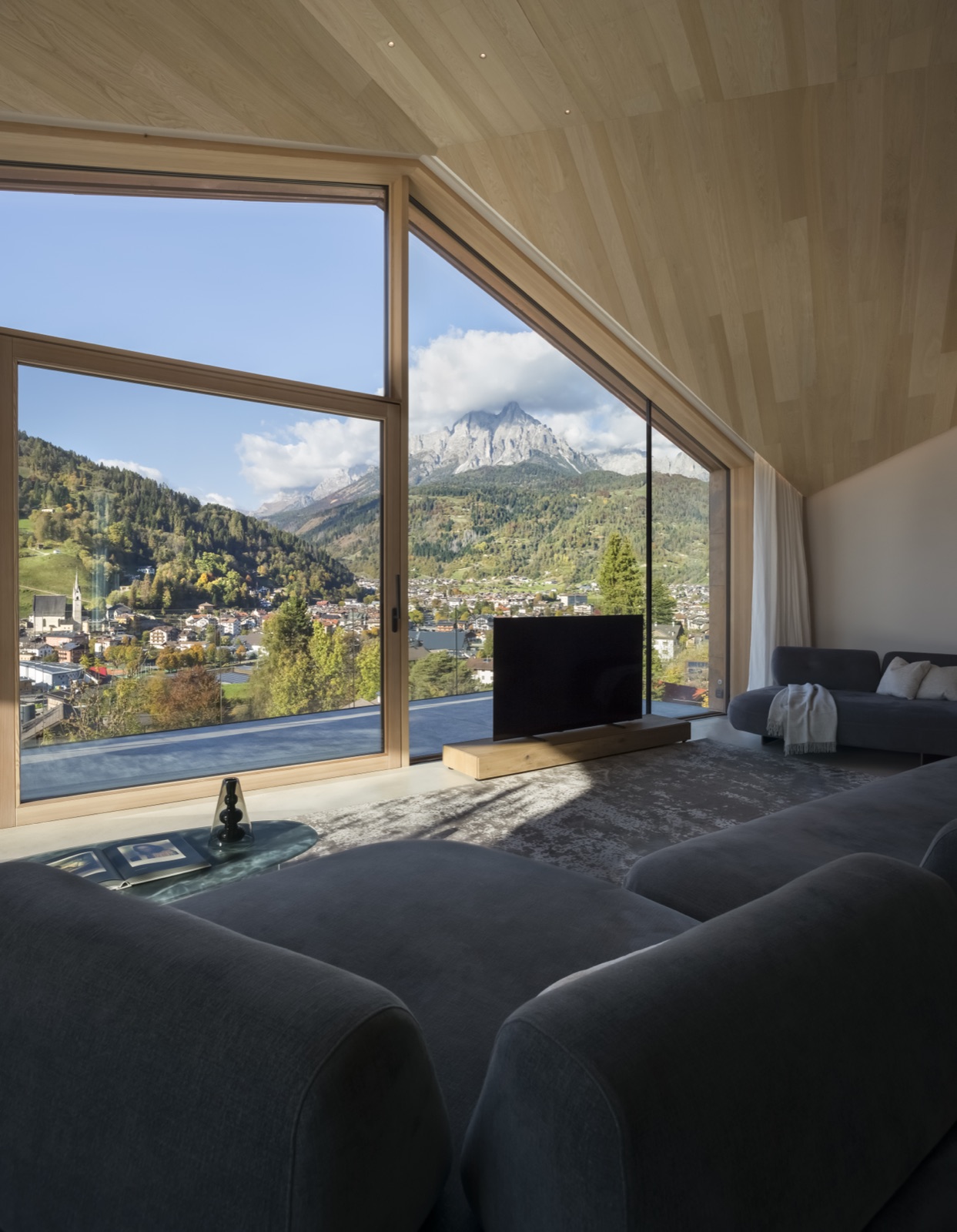 Le séjour de Casa LAGO Dolomiti, ses grandes baies vitrées ouvertes sur le paysage alpin et le village de Primiero
